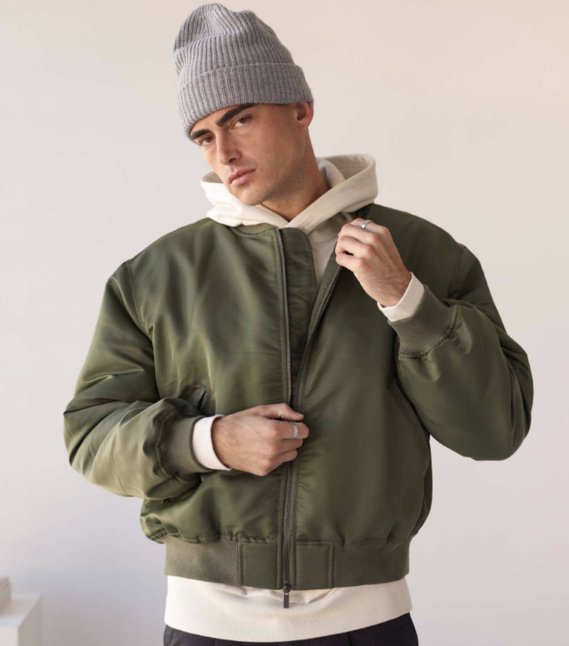 Model in an olive bomber jacket over a cream hoodie and grey beanie