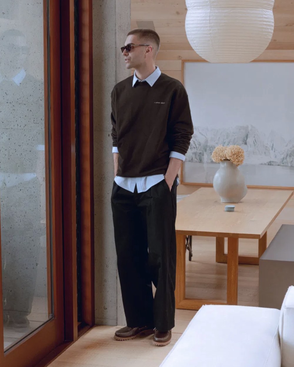 Model in a brown crewneck over a light blue shirt and black pleated trousers by a window