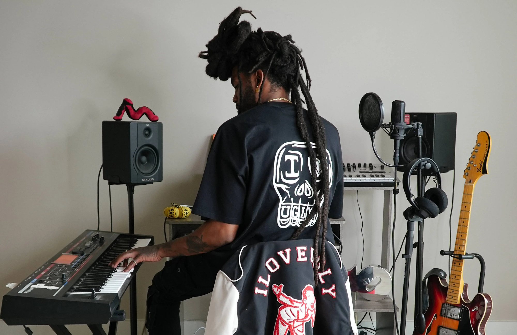 Musician at a keyboard in a studio wearing an I Love Ugly skull tee, with an embroidered varsity jacket