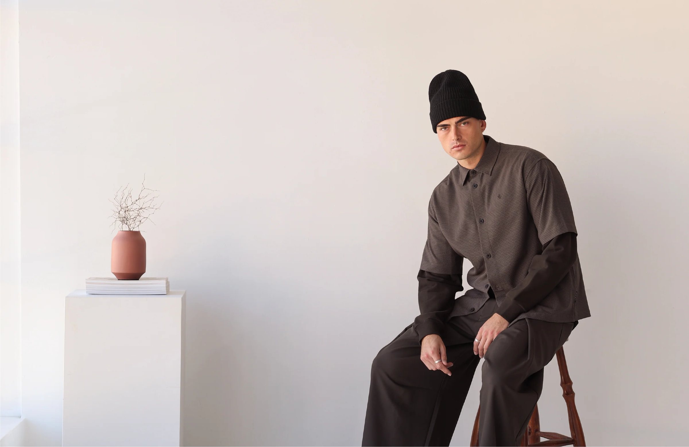 Model in a black beanie, textured short-sleeve shirt over a long-sleeve tee and wide trousers, seated on a stool beside a white plinth with a vase