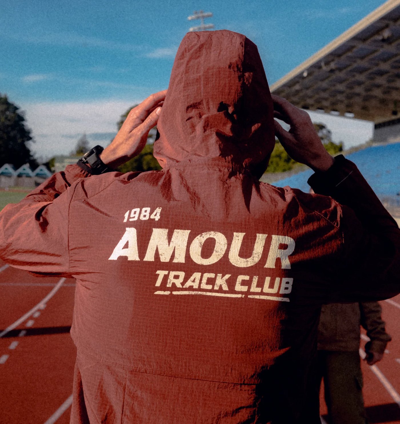 Rust ripstop windbreaker with large 1984 Amour Track Club back print, hood up on a running track