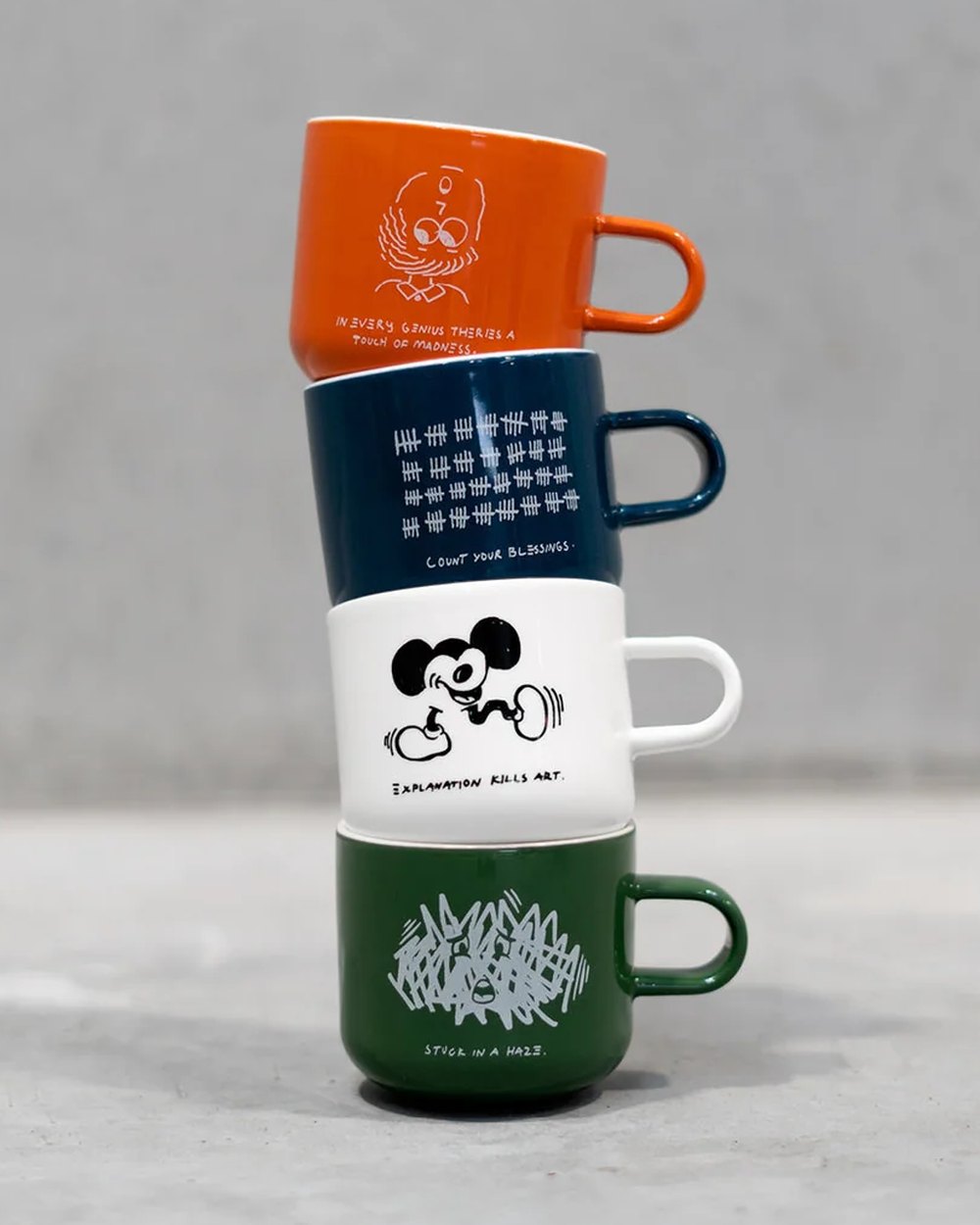 Stack of four ceramic mugs in orange, navy, white and green with illustrated quotes