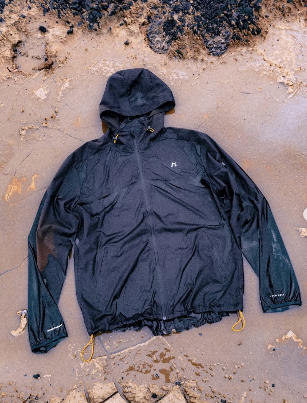 Black hooded running jacket with yellow toggles laid flat on wet sand