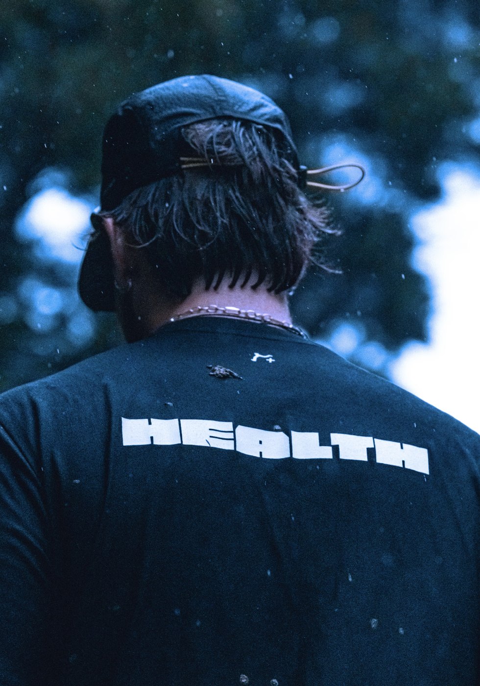 Back view of the black tee with HEALTH back print and five panel cap in the rain