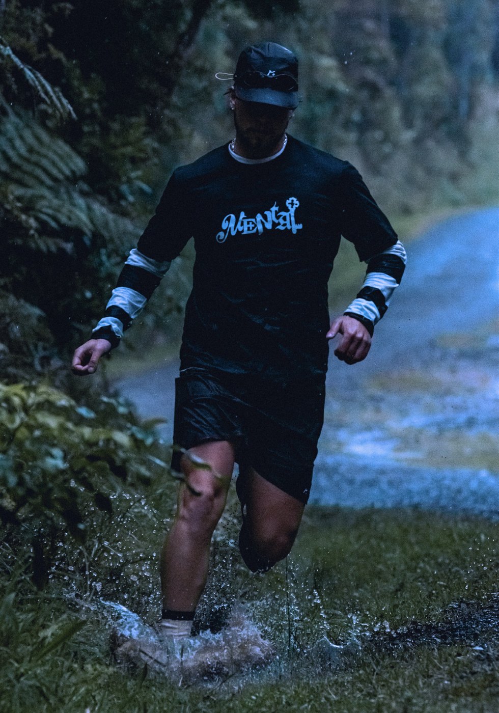 Runner splashing through a rainforest trail puddle in the black Mental tee with striped long sleeve layer